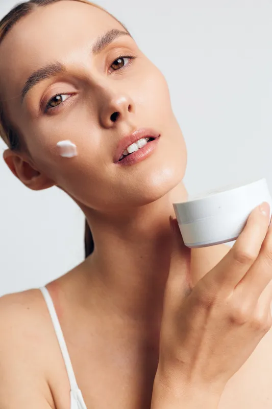 A young woman with clear skin applies white cream on her face holding a jar, posing against a plain light background. Concept for skincare, beauty, and facial treatment.