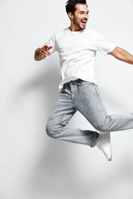 Happy joyful young man jumping in the air, wearing a casual white t-shirt and gray jeans, isolated on plain light background. People lifestyle concept.