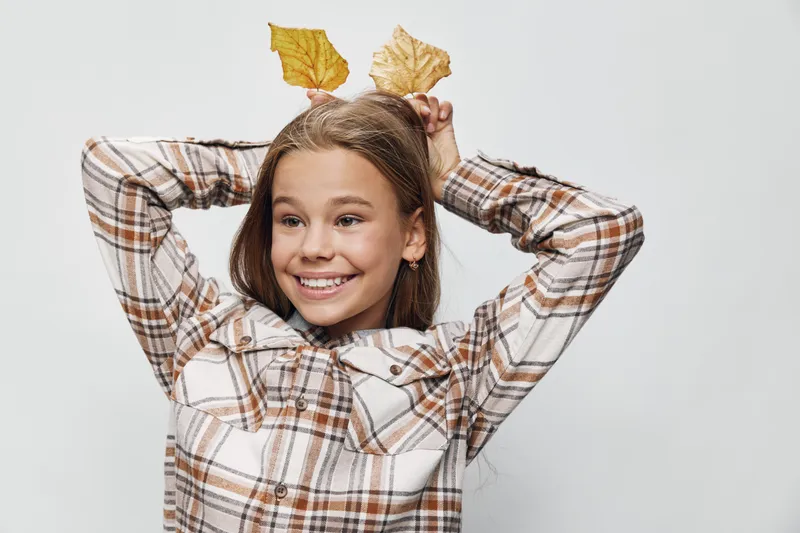 Girl smiling and posing with autumn leaves above head wearing plaid shirt in studio. Happy child with brown hair and casual warm outfit closeup on white background.