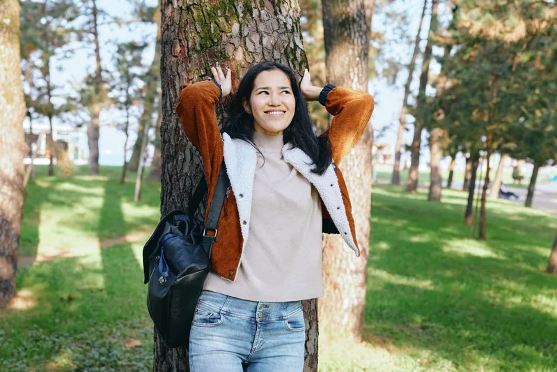 Woman outdoors park tree casual jacket with backpack smiling in sunlit greenery relaxed outdoor leisure moment amid nature and scenic landscape with breeze and daylight