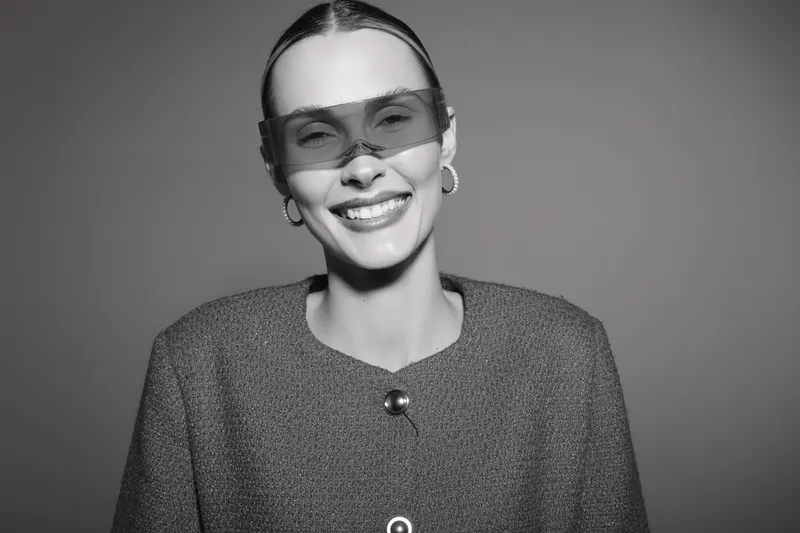 Smiling white woman with fair skin wearing stylish sunglasses and a textured jacket poses against a plain background. Concept of fashion, confidence, and modern style.