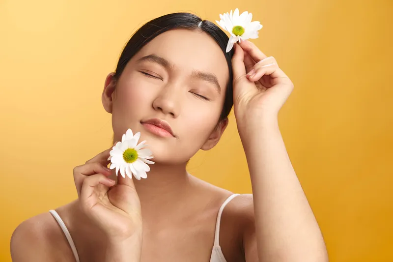 Asian woman gently holds flowers near her face in a soft studio portrait with a warm background, conveying calm beauty and a peaceful, graceful mood.