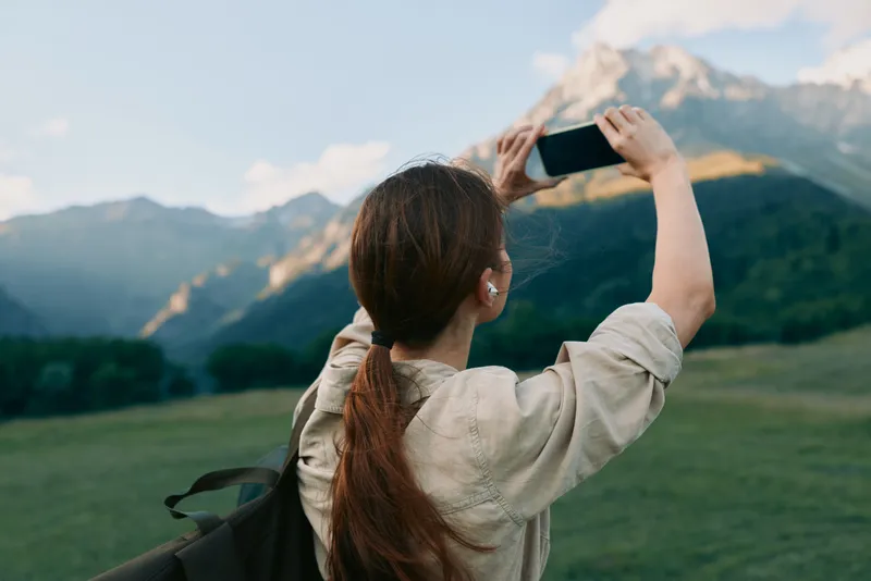 A young woman with a backpack takes a photo of distant mountains, capturing a moment of exploration and awe.