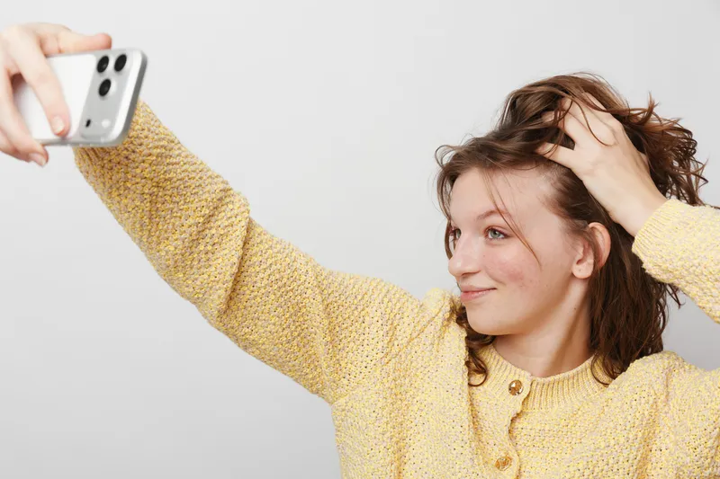 Young woman with curly hair wearing yellow sweater takes selfie with smartphone, smiling and touching hair. Studio portrait on white background. Concept of social media and self-expression.