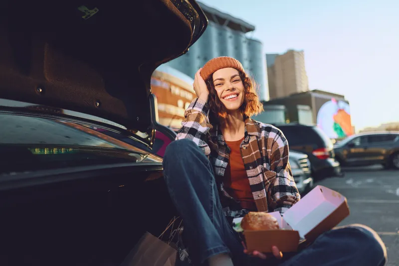 woman car food smile urban golden hour candid authenticity; young woman sits at open trunk eating from a takeaway box, joyful expression, golden hour glow, mindful living and emotional storytelling