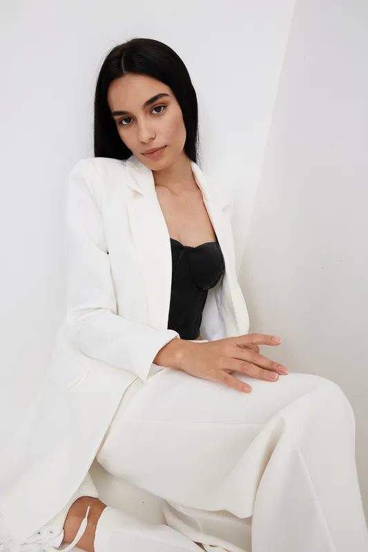 young woman in white suit sitting indoors with black top, dark hair, relaxed pose, minimalistic background, soft lighting, modern fashion style