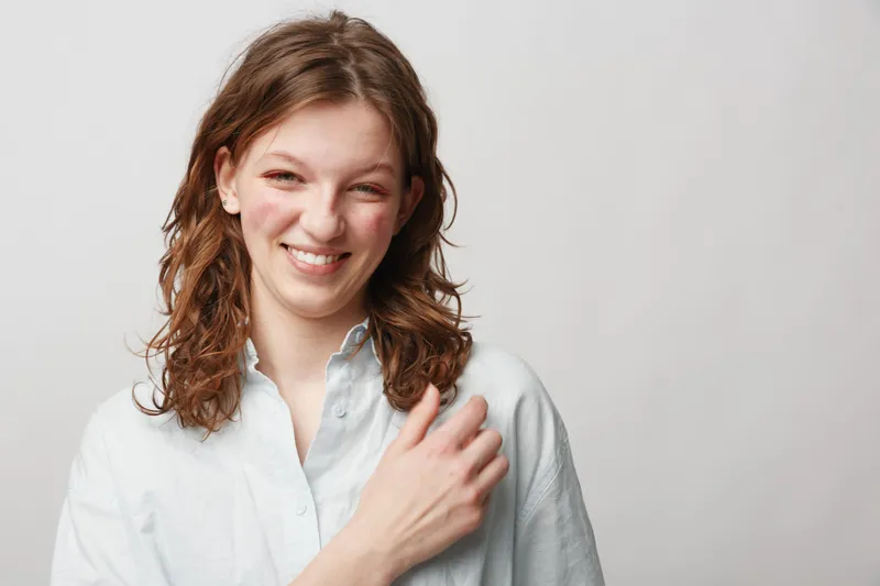 Smiling young woman with curly brown hair wearing white shirt looks happy and relaxed against plain light background. Concept of natural beauty, casual style, and positive emotions.