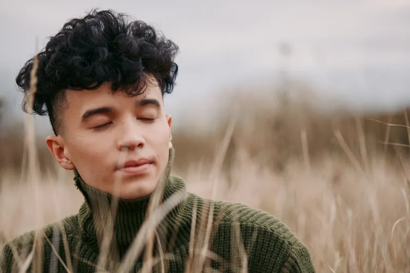 An authentic portrait highlighting credibility through a calm, grounded moment, as a youthful subject rests in a coarse knit sweater amid tall grass and soft light.