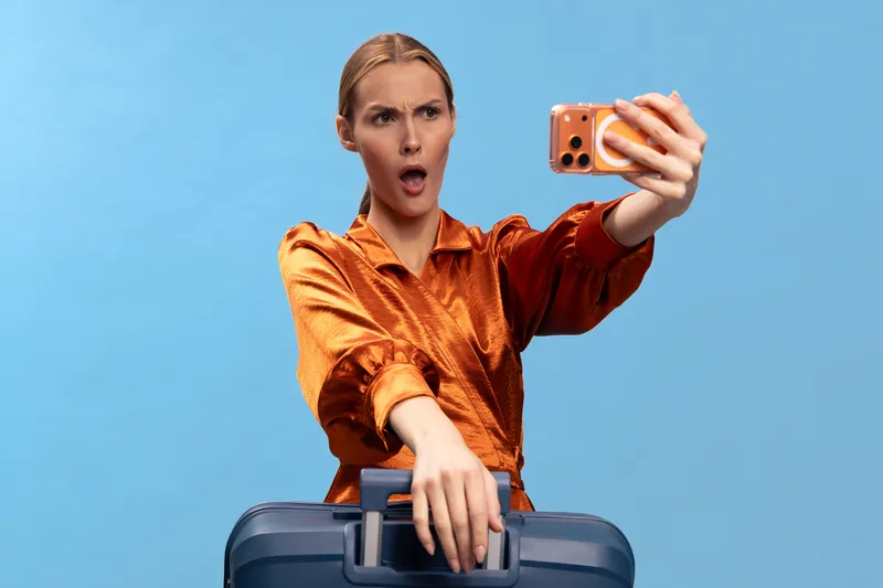 Surprised woman in orange satin blouse takes selfie with smartphone, holding navy suitcase handle. Blue background studio portrait, concept of travel, vacation, and unexpected moments.