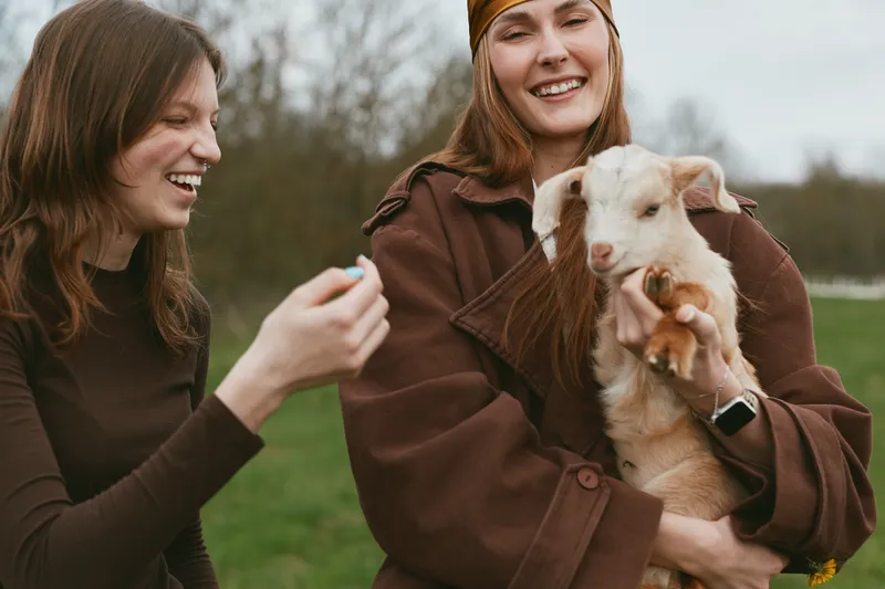 Goat kid is cradled by a smiling woman while another woman chats nearby in an outdoor setting, showcasing a warm lifestyle moment in a grassy field. Natural light emphasizes friendship, care, and