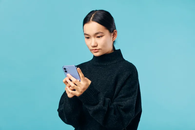 Woman wearing black sweater looking at smartphone with focused expression against blue background. Technology, communication, and casual modern lifestyle concept with mobile device.