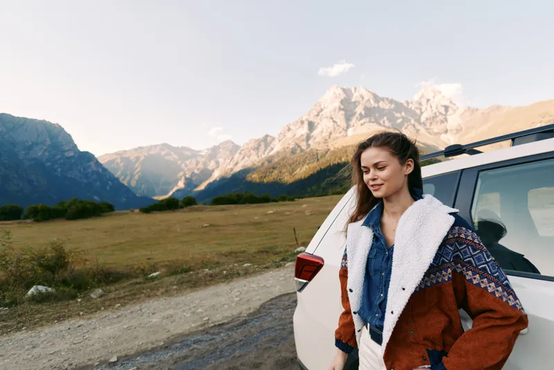 Woman by car on mountain road during outdoor travel, scenic landscape and nature view. Young female in warm jacket stands beside vehicle with peaks in background and calm expression