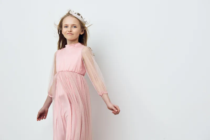 Elegant young girl in a light pink dress with a crown smiles softly, embodying innocence and joy against a minimalist white background
