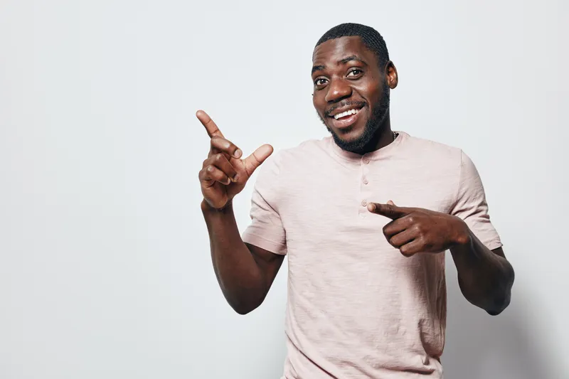 African American man with dark skin wearing light pink shirt smiling and pointing fingers on white background for casual lifestyle and positive emotion.
