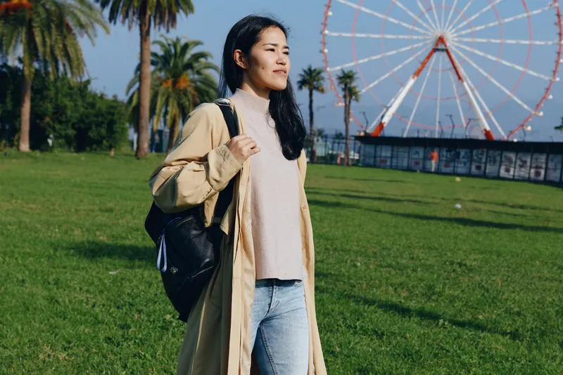 A woman with a backpack stands in a sunny park with a ferris wheel in the background, wearing a casual coat and jeans, enjoying outdoor leisure and travel vibes in a bright urban park setting