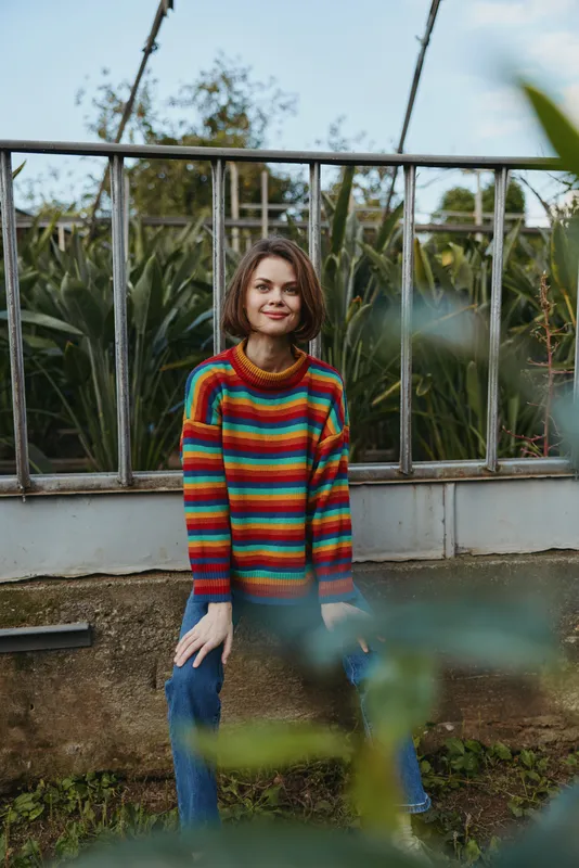Young woman wearing a rainbow striped sweater and blue jeans sitting on a low wall in a green garden, short hair smiling, casual outfit, relaxed outdoor lifestyle portrait