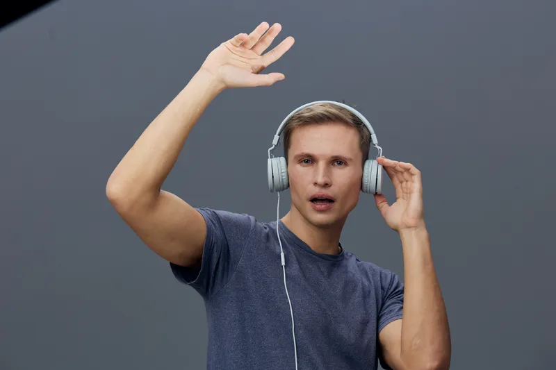 Young man with headphones listening to music on gray background, raising hand and singing with expression, casual clothing, studio portrait, people lifestyle concept.