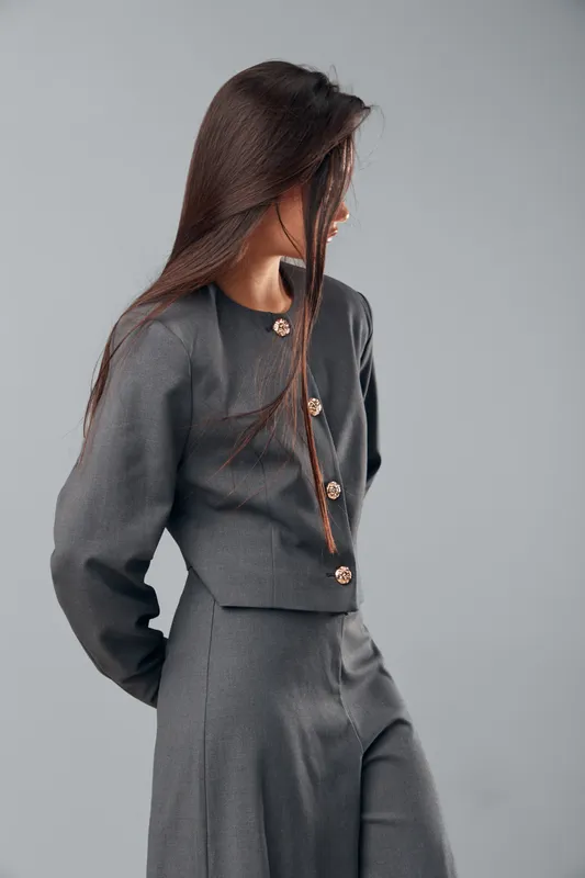 Woman wearing elegant gray suit with gold buttons poses sideways against gray background. Female model with long dark hair showcases outfit in fashion studio with minimalist style and confident