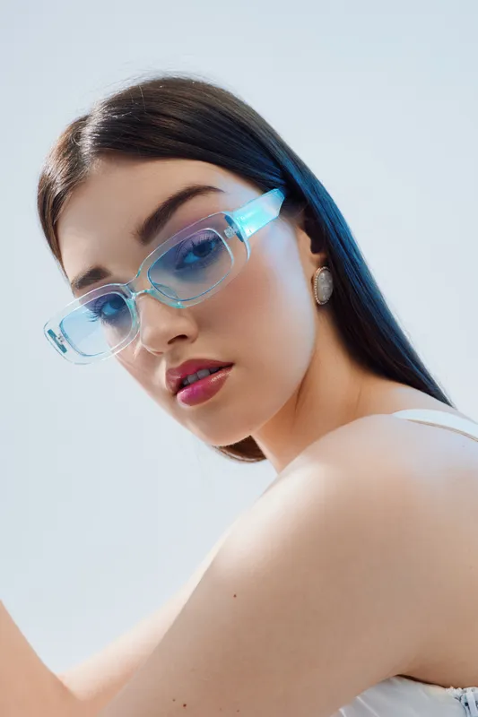 Young woman with long dark hair wearing transparent blue sunglasses and white top looks confidently at camera. Studio portrait with soft lighting. Concept of woman beauty fashion.