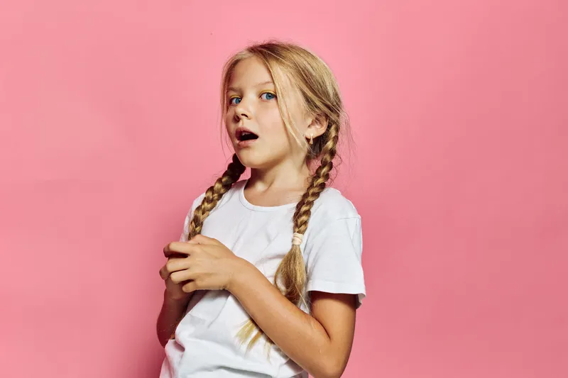 Surprised girl with braids and white t-shirt looks shocked on a pink background. Bright studio portrait concept of emotion.