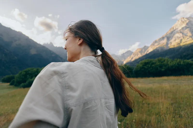 Young woman with long dark hair walks through a grassy field toward distant mountains, capturing a calm, adventurous outdoor mood as a gentle breeze rustles.