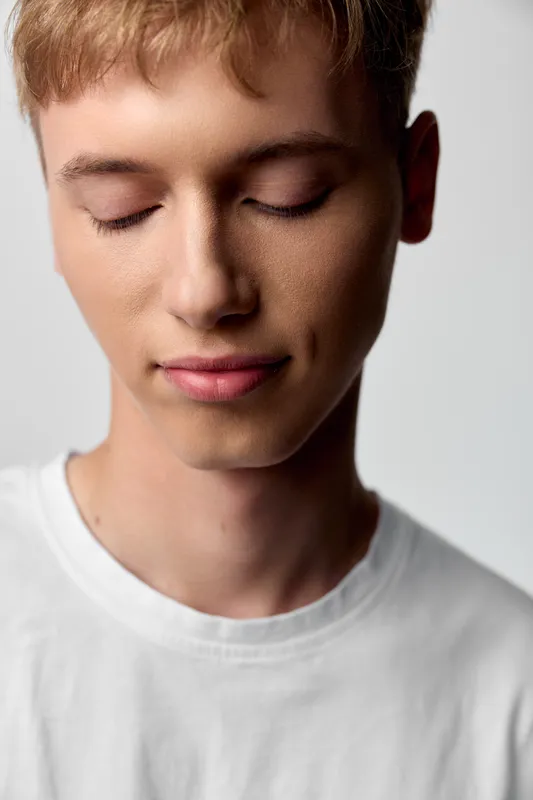 Young man with closed eyes calm expression soft lighting white shirt serene minimal studio mood emphasizing relaxation contemplation gentle beauty in a simple setup