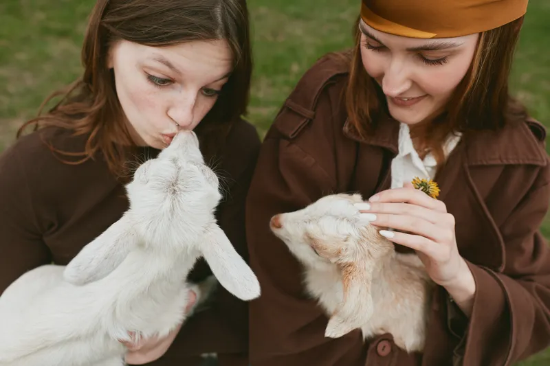 Two women in vintage outfits share a gentle moment with goats in an outdoor setting, capturing a lifestyle scene. Caring interaction, playful energy, and calm farm atmosphere with animal