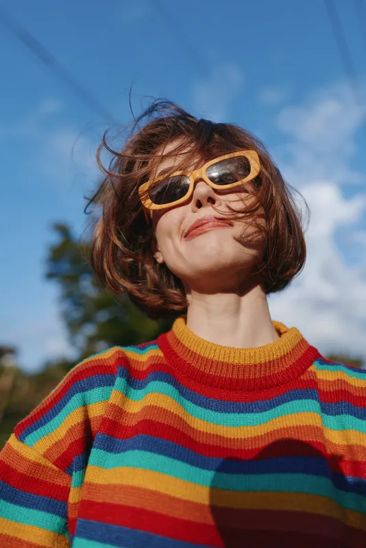 Woman wearing yellow sunglasses and a colorful striped sweater, smiling with short bob haircut outdoors under blue sky. Mid 30s model, playful windblown hair, retro casual outfit for lifestyle and