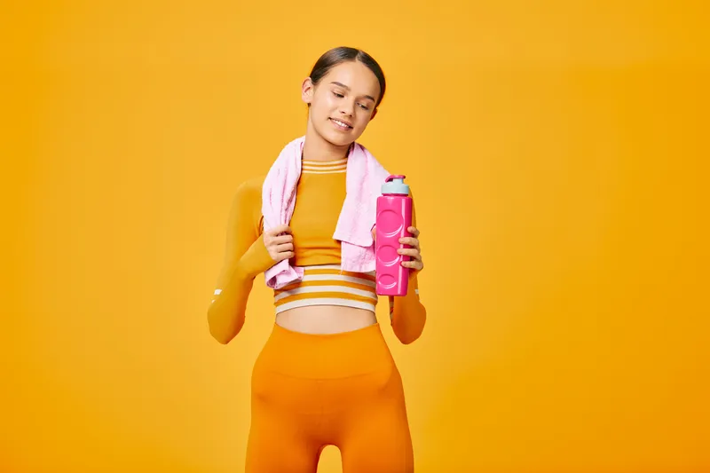 Sporty woman in orange athletic wear with a pink water bottle and a towel around her neck, posing confidently in a vivid orange studio after a workout.