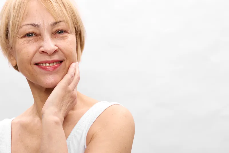 Smiling senior blonde woman in studio close up, touching her cheek while using skincare on her face, bright white background, conveying wellness and health, gentle mood