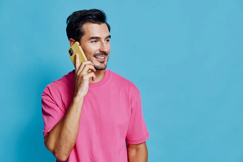 Smiling man wearing a pink shirt holds a mobile phone, looking cheerful against a vibrant blue background. People lifestyle concept.