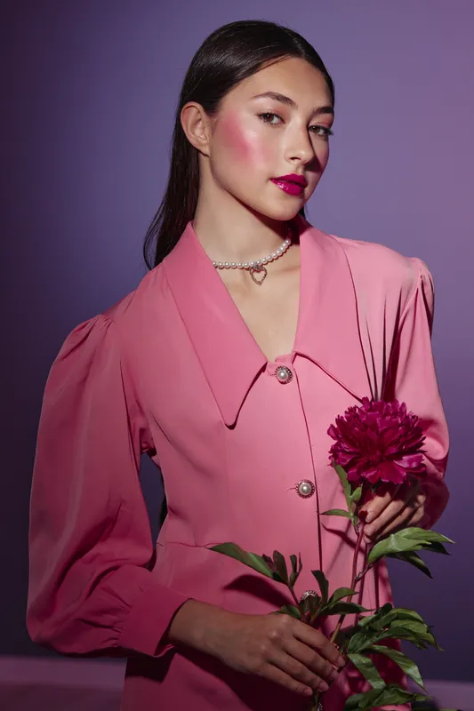 Young elegant woman with model makeup wears stylish pink outfit and pearl necklace while holding a large dark pink flower. Glamorous fashion beauty concept on purple background.