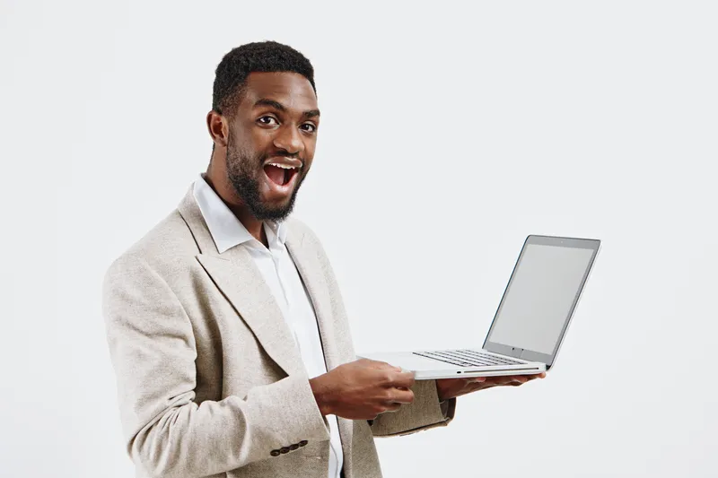 Cheerful young man with dark skin and short curly hair holding a laptop computer in his right hand, dressed in a beige textured blazer and white shirt, looking excited and happy, standing against a pl