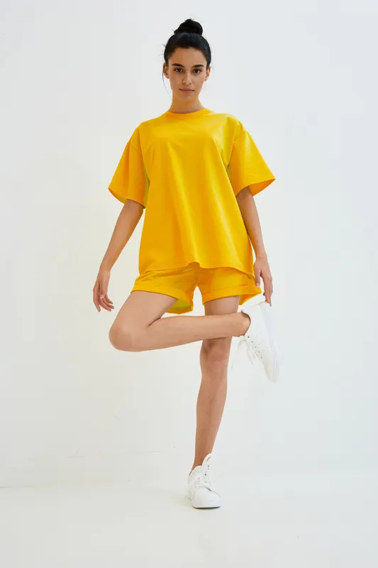 Young woman standing on one leg wearing bright yellow casual outfit and white sneakers on plain white background, showcasing relaxed summer style and sporty vibe.