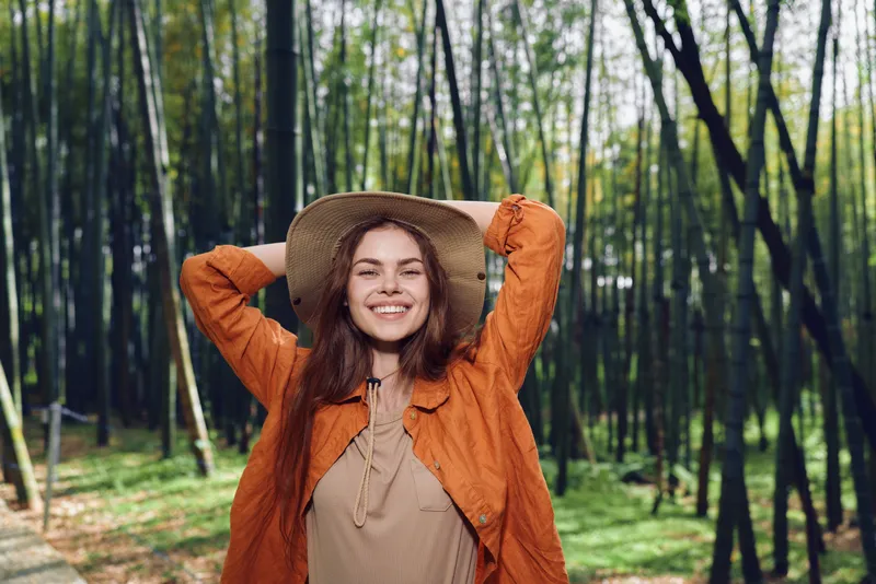 Woman smile hat forest bamboo outdoors nature portrait of a young woman in a straw hat and orange jacket, hands behind head, joyful expression during a sunny walk in woodland.