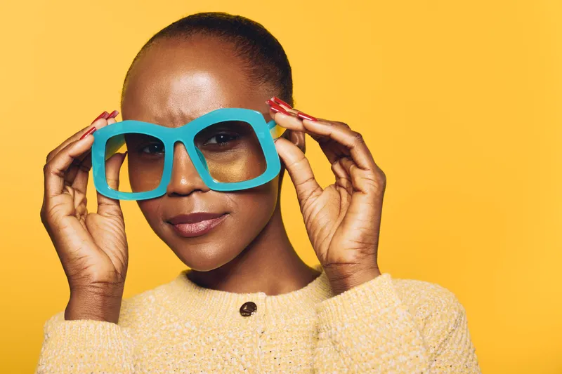 Close-up of confident African woman with dark skin wearing oversized blue glasses and yellow sweater. Studio portrait on yellow background. Concept of fashion, style, and beauty.