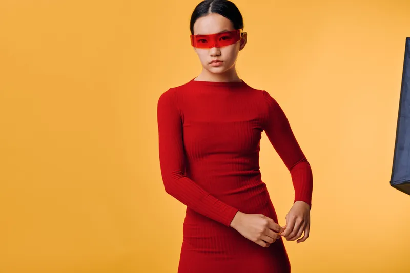 Young woman in red fitted dress and red tinted sunglasses posing against yellow background with stylish hair tied back and confident expression in studio fashion portrait