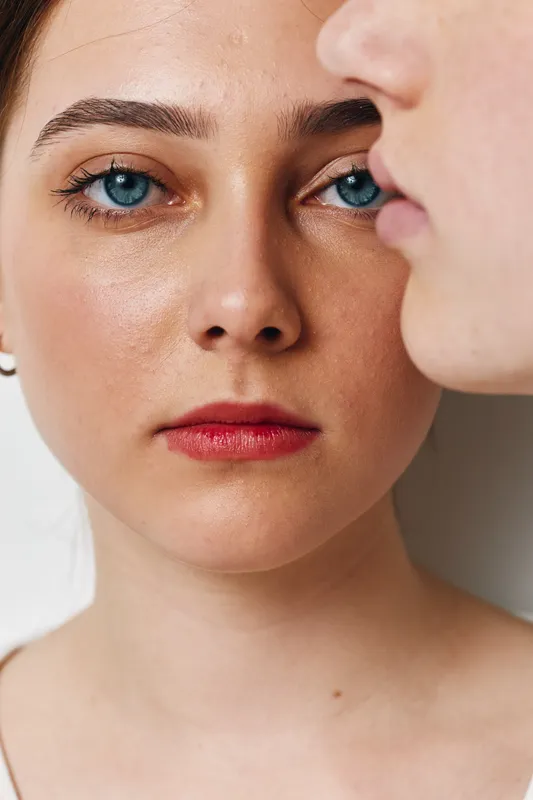 Closeup of two young women faces with natural skin and blue eyes against white background, showing emotions and intimate portrait with detailed texture and makeup.