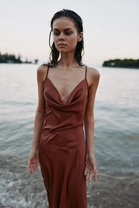Portrait of a woman standing by a calm shoreline wearing a rust brown satin slip dress, wet hair, soft light, and thoughtful expression, capturing calm coastal mood against evening glow and gentle
