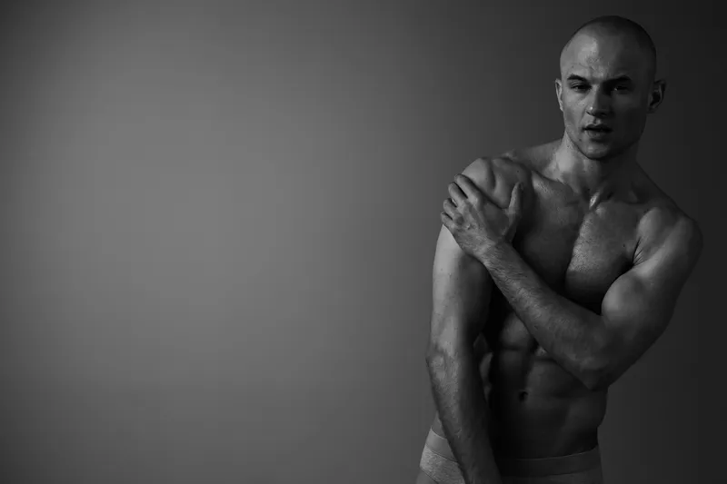 A striking black and white portrait of a man showcasing his toned physique with a dramatic pose, capturing strength and vulnerability in a minimalistic setting.