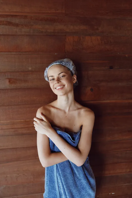 Smiling woman wrapped in blue towel with headscarf, standing against wooden wall, expressing relaxation and natural beauty in warm indoor spa environment.