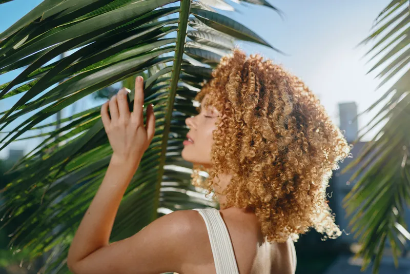 Curly woman with natural hair enjoying sunlight behind green palm leaves, wearing white sleeveless top, warm outdoor ambiance, summer feel with soft focus.