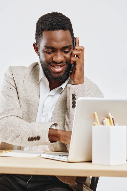 Smiling man talking on mobile phone, working at desk with laptop, white background, casual business attire, modern workspace, cheerful mood, communication concept, adult male, Africa, technology, busi