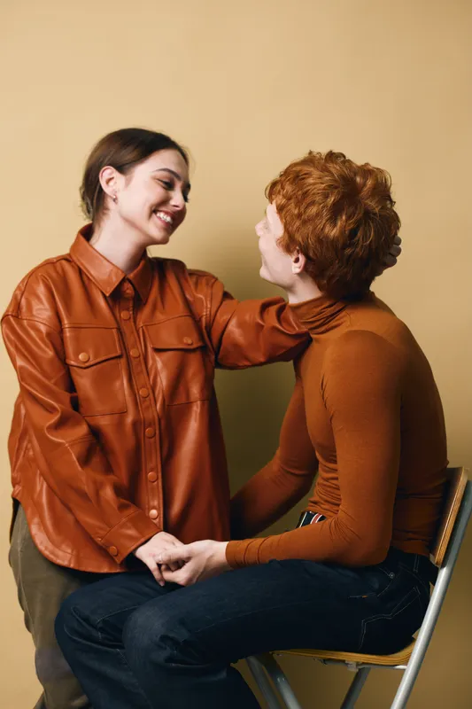 Couple sitting and smiling at each other, holding hands with casual autumn clothing indoors. Young woman and man showing affection and connection in relationships.