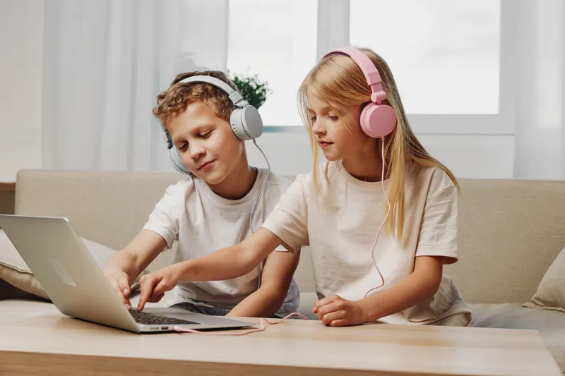 Two kids in headphones sit on sofa using laptop together in bright room. Concept of online learning, technology, children teamwork, education, and digital communication at home environment.