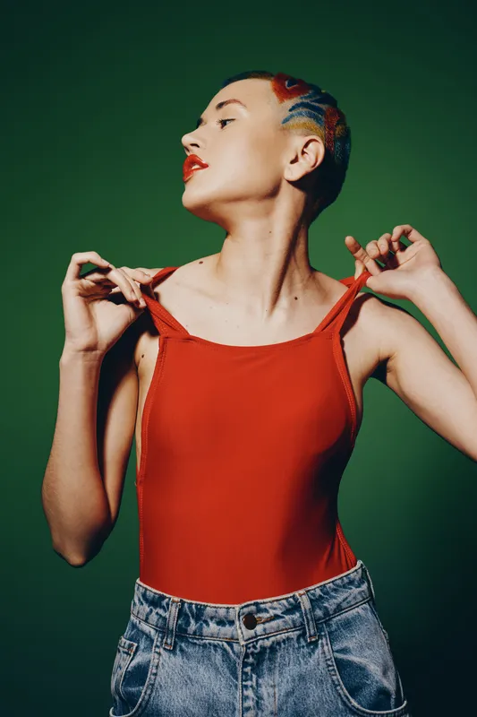 Woman with colorful braided hair wearing red top and denim jeans poses confidently against green background, displaying unique style and bold expression in studio lighting.