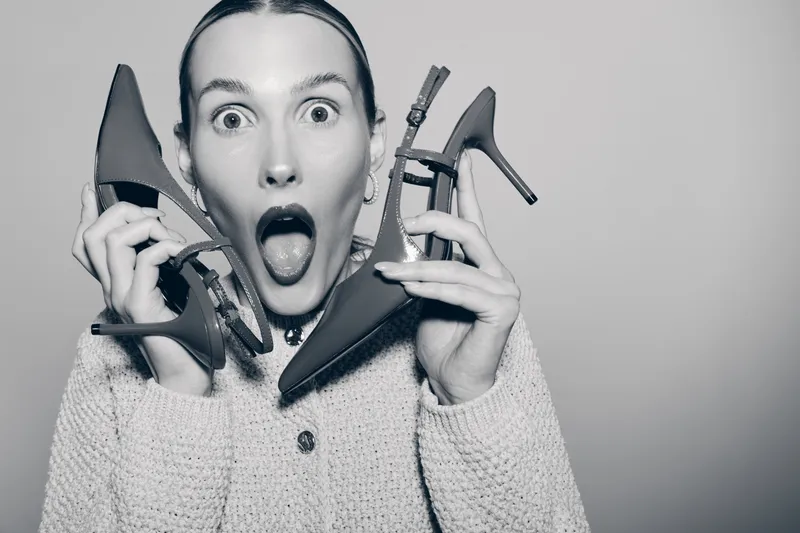 White woman with light skin holds two high heel shoes near her face with surprised expression on grey background.