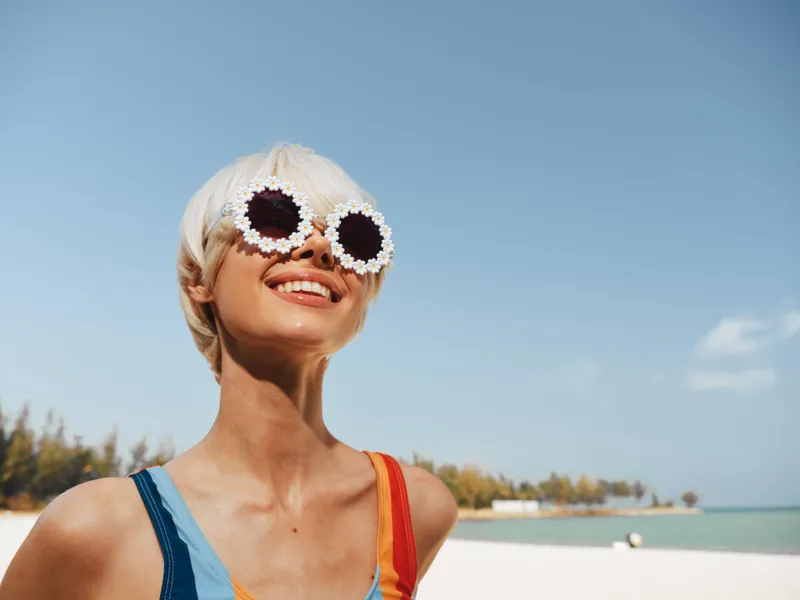 A joyful woman at the beach wears crochet sunglasses and colorful straps, capturing a sunny, carefree summer portrait with a bright sky and distant shoreline.