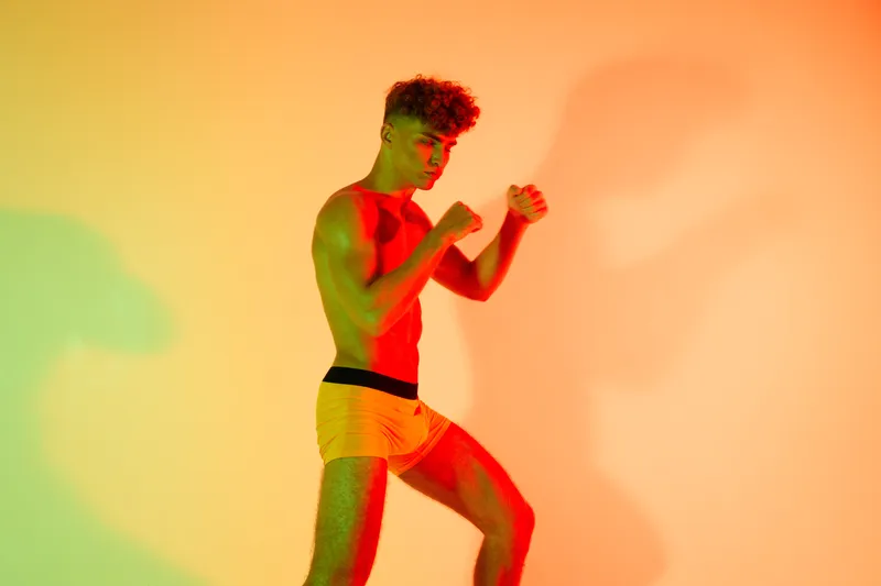 Young man in yellow shorts posing in fighting stance with colorful lighting and shadows on orange background for fitness and boxing concept.