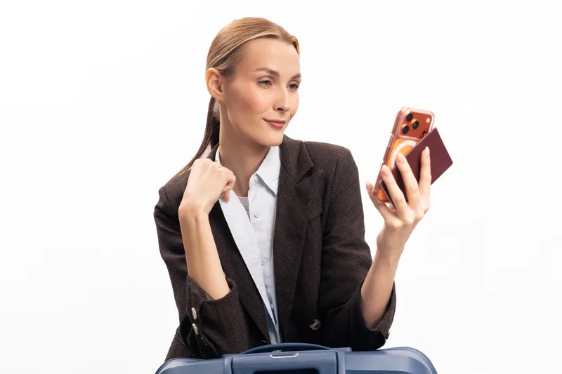 Confident businesswoman holds passport and smartphone with orange case, standing behind blue suitcase. Concept for travel, business trip, mobile communication, and modern lifestyle.